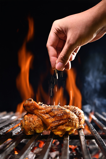 Hand sprinkling salt and spices on grilled chicken leg on the flaming grill