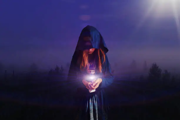 Photo of A girl with a hood on her head walks through the night forest and carries a lamp. The light spreads and illuminates the night. The concept of prayer, holiness, mysticism, magic.