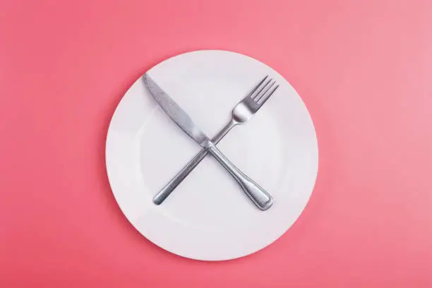 Photo of Empty plate on a pink minimal background. Empty white ceramic plate with knife and fork on the table after eating. Diet and healthy food concept.