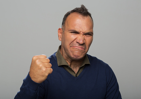 Angry mature man with eyes closed showing fist standing against grey background.