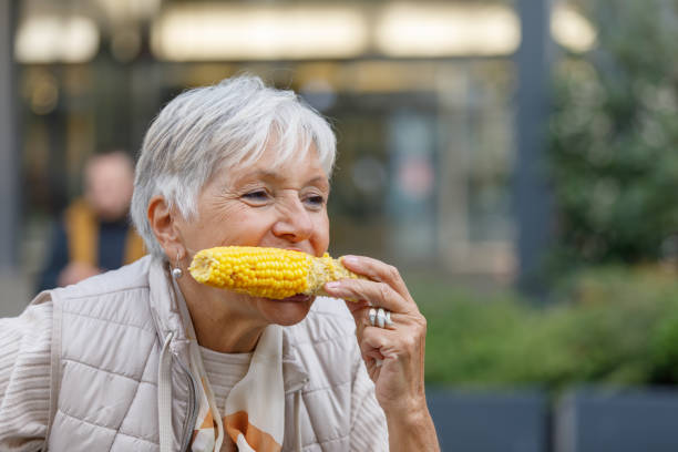 uma mulher mais velha está comendo uma espiga de milho cozido e caminhando na cidade. - corn on the cob fotos - fotografias e filmes do acervo