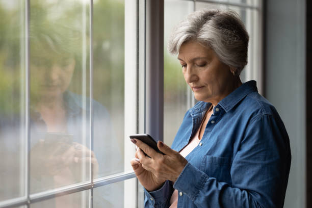 istock Serious latin female pensioner stand by window texting by cell 1311594944