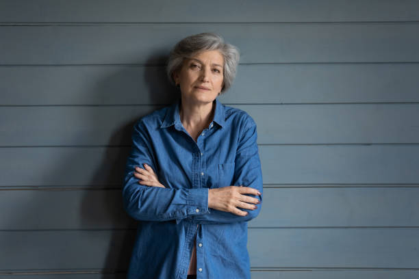 Serious elderly latin lady posing for portrait keep arms crossed Charismatic person. Serious elderly latin lady posing for portrait keep arms crossed. Confident old female mother grandma in stylish jeans jacket stand by grey planked wall look at camera. Copy space grandma portrait stock pictures, royalty-free photos & images