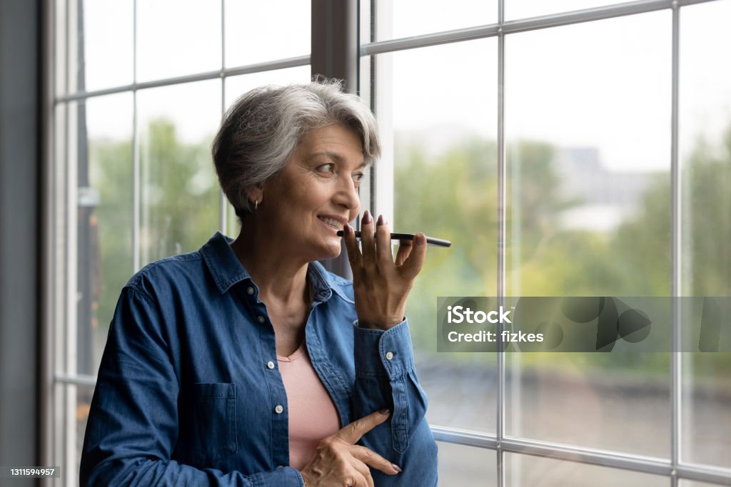 Confident aged hispanic female hold modern smartphone dictate audio message Easy communication. Confident aged hispanic female look at large window hold modern smartphone dictate audio message. Smiling senior latina woman give vocal command to digital ai assistant. Copy space Conference Phone Stock Photo