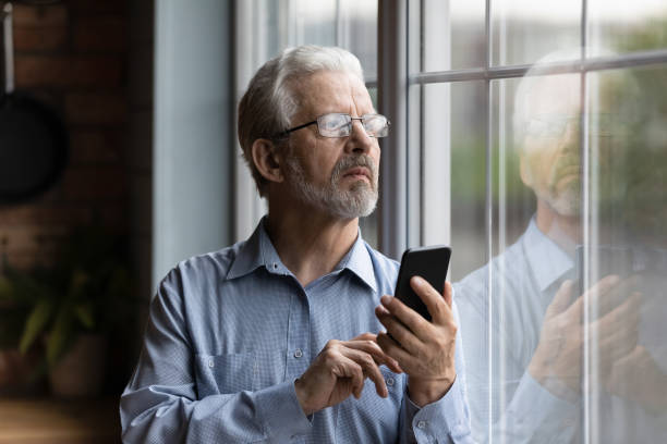 istock Worried older man look at window hold phone make call 1311595007