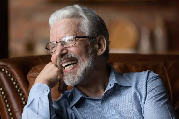 Mature man sit on sofa laugh demonstrate healthy straight teeth Happy life on pension. Mature aged man retiree wear glasses sit on sofa look aside laugh demonstrate healthy straight teeth prothesis implants. Old man with white smile advertise dental clinic service dentures stock pictures, royalty-free photos & images