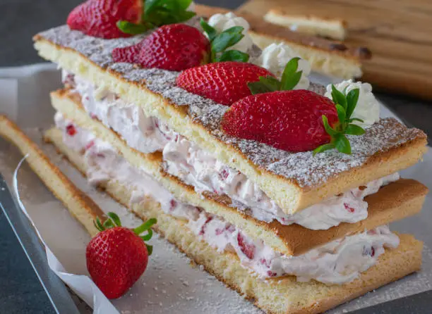 Delicious homemade strawberry cream cake served as a square layer cake with whipped cream and fresh strawberries cubes on a table. Ready to eat