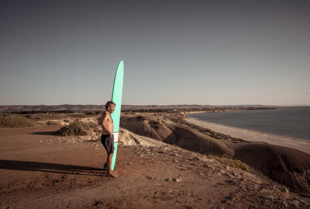 attraente surfista australiano maturo con tavola da surf vintage fresca sulla spiaggia al tramonto. adulto anziano felice di essere tornato a surfare. avventura sportiva all'aria aperta, anziani attivi e stile di vita pensionistico. - senior adult surfing aging process sport foto e immagini stock