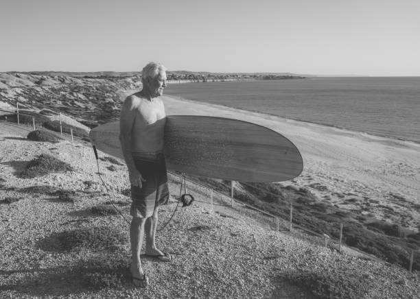 attraente surfista australiano maturo con tavola da surf vintage fresca sulla spiaggia al tramonto. adulto anziano felice di essere tornato a surfare. avventura sportiva all'aria aperta, anziani attivi e stile di vita pensionistico. - senior adult surfing aging process sport foto e immagini stock