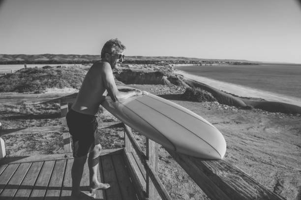 attraente surfista australiano maturo con tavola da surf vintage fresca sulla spiaggia al tramonto. adulto anziano felice di essere tornato a surfare. avventura sportiva all'aria aperta, anziani attivi e stile di vita pensionistico. - senior adult surfing aging process sport foto e immagini stock