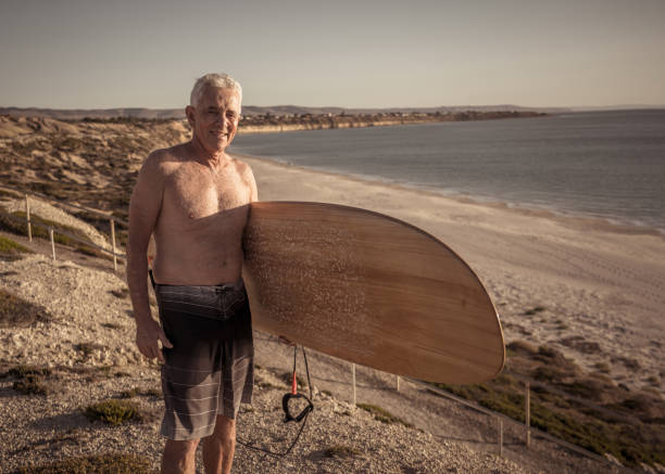 surfer mûr attirant d’homme avec la planche de surf vintage fraîche sur la plage au coucher du soleil. adulte aîné heureux d’être de retour pour surfer. aventure sportive en plein air, personnes âgées actives et mode de vie à la retraite. - senior adult surfing aging process sport photos et images de collection