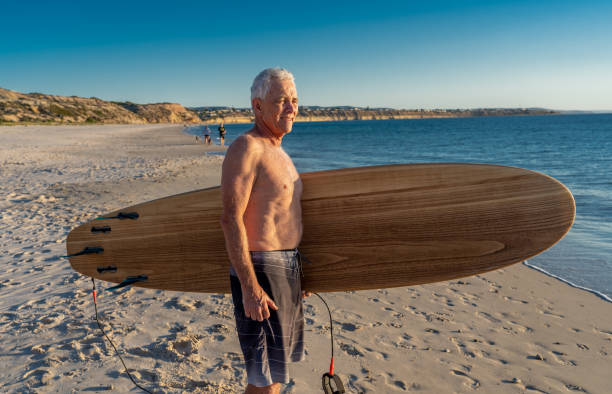 attraente surfista australiano maturo con tavola da surf vintage fresca sulla spiaggia al tramonto. adulto anziano felice di essere tornato a surfare. avventura sportiva all'aria aperta, anziani attivi e stile di vita pensionistico. - senior adult surfing aging process sport foto e immagini stock