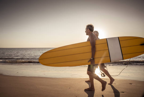 deux hommes mûrs marchant avec des planches de surf sur la belle plage appréciant le paradis et le mode de vie de retraite. attrayant ajustement amis adultes seniors ayant du plaisir à surfer. chez les personnes actives et en bonne santé. - senior adult surfing aging process sport photos et images de collection