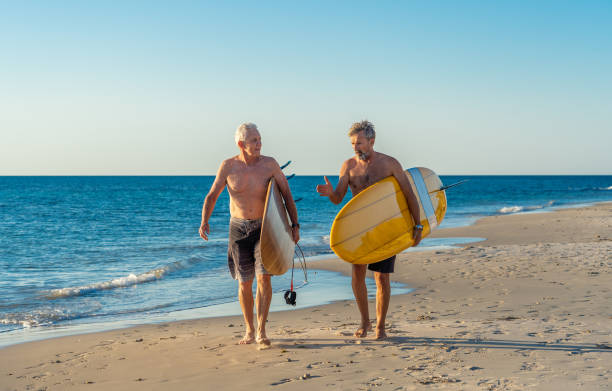 deux hommes mûrs marchant avec des planches de surf sur la belle plage appréciant le paradis et le mode de vie de retraite. attrayant ajustement amis adultes seniors ayant du plaisir à surfer. chez les personnes actives et en bonne santé. - senior adult surfing aging process sport photos et images de collection