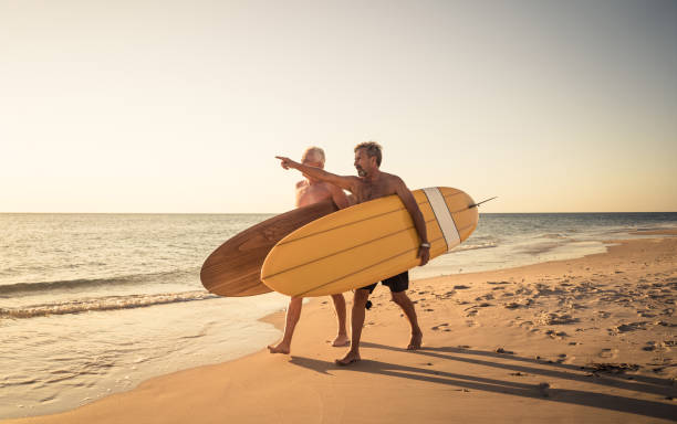 deux hommes mûrs marchant avec des planches de surf sur la belle plage appréciant le paradis et le mode de vie de retraite. attrayant ajustement amis adultes seniors ayant du plaisir à surfer. chez les personnes actives et en bonne santé. - senior adult surfing aging process sport photos et images de collection