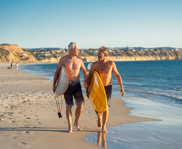 due uomini maturi che camminano con tavole da surf sulla bellissima spiaggia godendo del paradiso e dello stile di vita pensionistico. attraente vestibilità amici adulti anziani che si divertono a navigare. in persone reali attive e sane. - senior adult surfing aging process sport foto e immagini stock