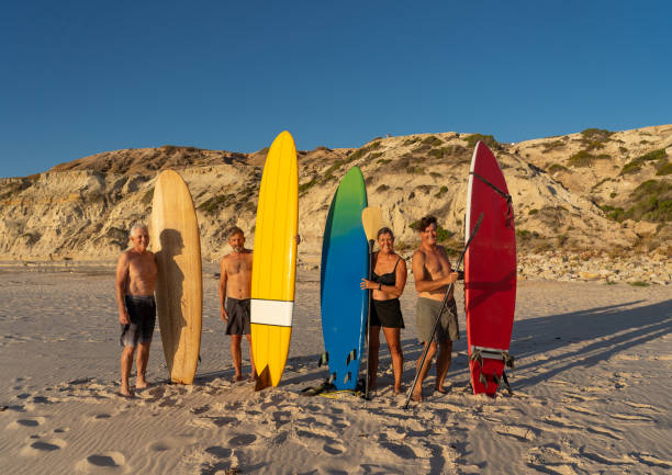 groupe de surfers aînés, femme et hommes, retenant leurs planches colorées de surf sur la plage éloignée. amis retraités mûrs appréciant le surf et le mode de vie extérieur dans les personnes en bonne santé et vieillissant dans le monde moderne. - senior adult surfing aging process sport photos et images de collection