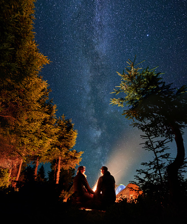 Young man and woman travelers sitting near bonfire under beautiful night sky with stars and Milky Way. Picturesque view of night starry sky over mountain forest with trees and hikers.