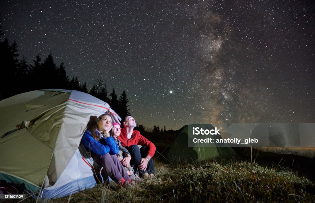 Travelers sitting in camp tent under night starry sky. Family hikers sitting in tourist tent under beautiful night sky with stars. Travelers resting in camp tent and looking at magical starry sky with Milky Way. Night camping in the mountains. Camping Stock Photo