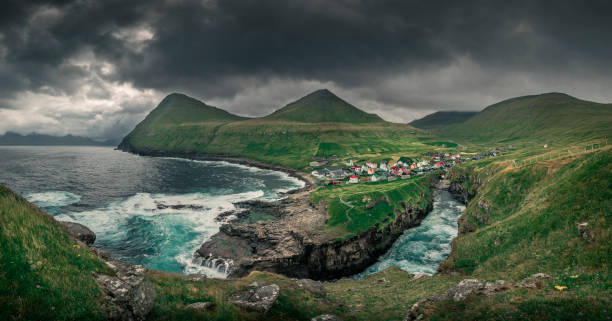 Panorama of gorge with the village of Gjogv on Eysturoy with green grass in front and mountains in back, Faroe Islands Panorama view of the gorge in the village of Gjogv on Eysturoy with green grass in front and mountains in back, Faroe Islands, ocean and dark cloudy dramatic sky eysturoy photos stock pictures, royalty-free photos & images