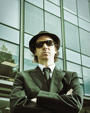 Serious, intimidating man wearing a suit, fedora hat and sunglasses glares down at you with folded arms. Copy space on the building behind him.