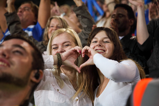 Two caucasian young women making heart with hands in a crowd on a stadium.