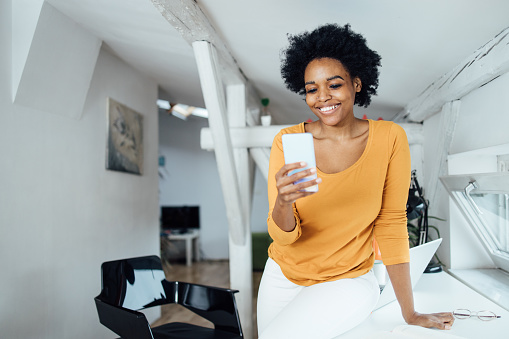 Portrait of a beautiful African-American woman using a smartphone and enjoying at her home office.