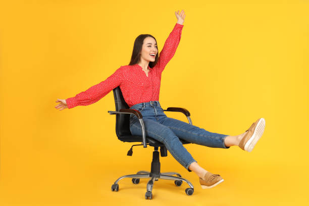 Young woman riding office chair on yellow background Young woman riding office chair on yellow background office chair stock pictures, royalty-free photos & images