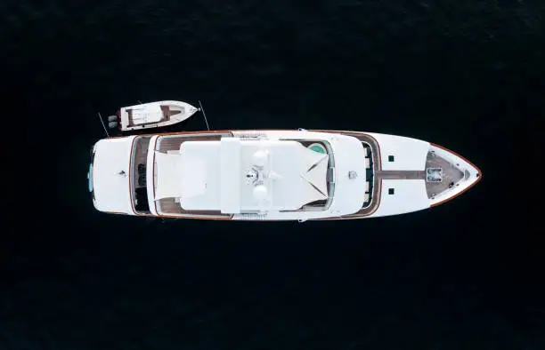 High angle aerial bird's eye view of a long white luxury yacht with wooden deck, a hot tub and a dinghy or tender boat on the side in dark blue waters shortly after sunset in Sydney, Australia.