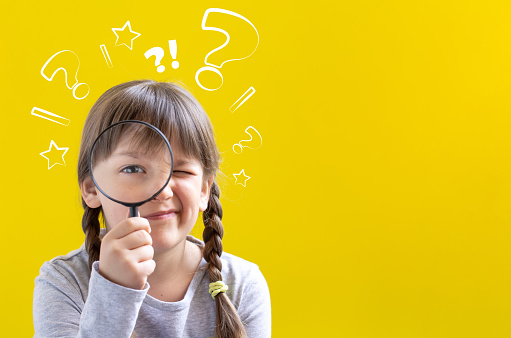 Cute curious little girl looking through a magnifying glass on yellow background with copy space.