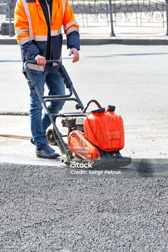 A road worker in an orange jacket compacts a patch of asphalt with a petrol vibrator. A road worker in a bright orange reflective jacket uses a vibratory plate to compact the asphalt at a road repair site. Vertical image, copy space. Compactor Stock Photo