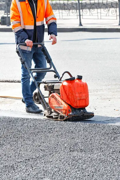 A road worker in a bright orange reflective jacket uses a vibratory plate to compact the asphalt at a road repair site. Vertical image, copy space.
