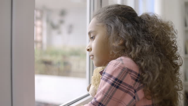Sad little girl looking through window, hugging favorite toy, waiting adoption