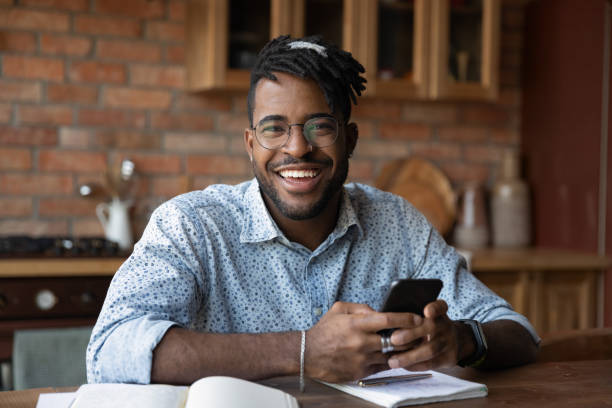 Portrait of successful black male modern day student holding smartphone Portrait of successful black male modern day student distracted from working learning look at camera hold smartphone. Happy smiling young afro american man hipster posing at kitchen table do paperwork networking photos stock pictures, royalty-free photos & images