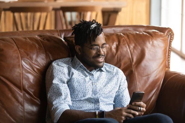 Joyful millennial african guy student play smartphone based online game Passionate gamer. Joyful millennial african guy student relax at home sit on couch play smartphone based online game. Young black man hipster addicted to internet spend free time using mobile phone image based social media photos stock pictures, royalty-free photos & images