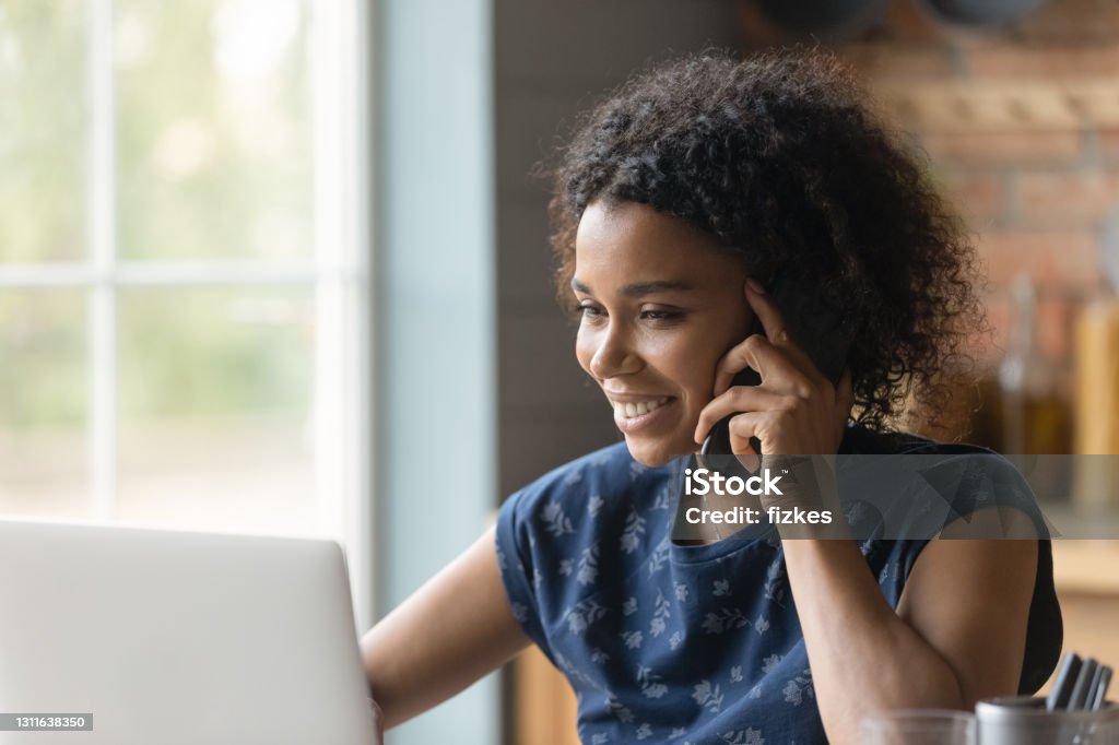 Biracial female hold cell at ear look at pc screen Modern way of communication. Smiling biracial female hold cell at ear talk look at pc screen. Busy young black lady advisor lawyer give phone consultation from home discuss electronic document details Using Phone Stock Photo