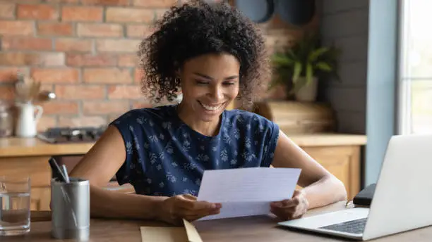 Happy smiling remote working young black female receive postal letter from company office about project report approval. Glad millennial afro american employee reading good news from paper document