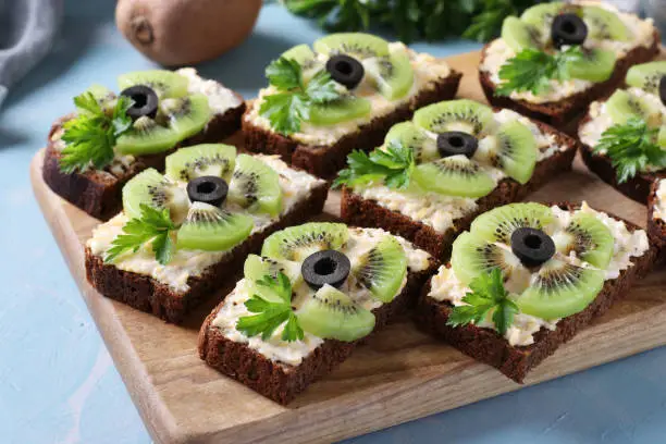 Homemade sandwiches with kiwi, cheese, garlic and black olives on wooden board on light blue background.