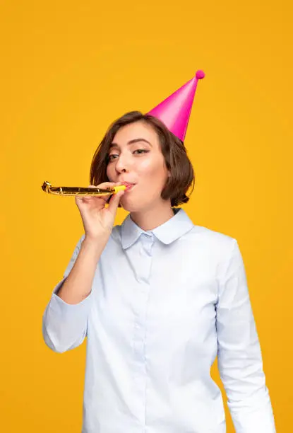 Young female in shirt and pink party hat looking away and blowing whistle while celebrating birthday against yellow background