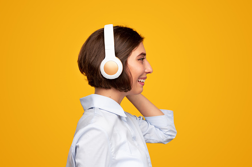 Cheerful young female meloman in casual clothes and wireless headphones listening to good music and smiling while standing against bright yellow background