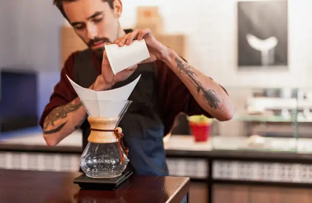 Concentrated male barista in apron standing at counter in coffee shop and making delicious beverage in glass coffeemaker