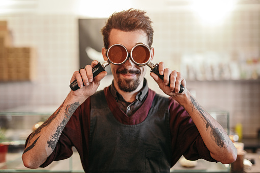 Positive bearded male bartender with tattooed arms covering eyes with portafilters while working in modern coffee shop