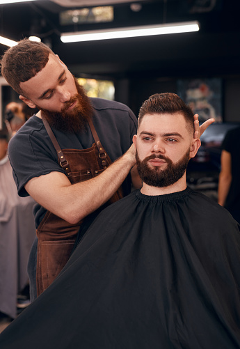 Man in apron demonstrating trendy haircut to bearded male customer while working in modern barbershop