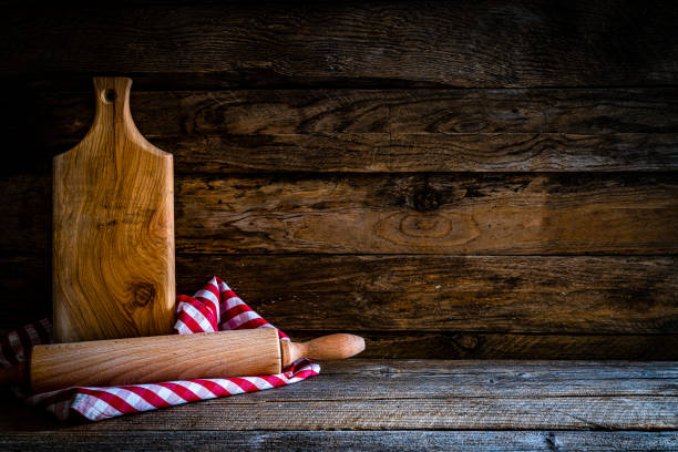 Cooking or kitchen backgrounds: front view of a an old cutting board and a rolling pin shot on rustic kitchen table. The cutting board and the rolling pin are on a crumpled gingham tablecloth. The composition is at the left of an horizontal frame leaving useful copy space for text and/or logo at the right. Predominant color is brown. High resolution 42Mp studio digital capture taken with SONY A7rII and Zeiss Batis 40mm F2.0 CF lens