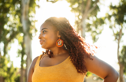 Confident young African woman glancing sideways and smiling while standing outside in a park on a sunny summer afternoon