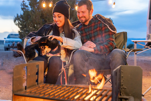 Smiling Young Couple Staying Warm By The Campfire, The Young Woman Playfully Petting Their Dog