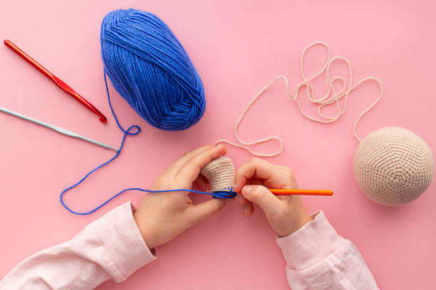 Children's hands in the process of crocheting toys from blue and beige yarn. Children's hands in the process of crocheting toys from blue and beige yarn. Pink background. Needlework, hobbies, craft training. The development of fine motor skills. Knitting school. Flat lay. Crochet stock pictures, royalty-free photos & images