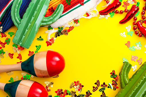 Mexican fiesta and Cinco de Mayo party concept theme with jalapeno pepper necklace, maracas, cactus and traditional rug covered in sombrero shaped confetti on yellow background with copy space
