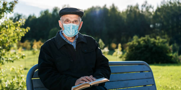 istock Elder man wearing eyewear and facemask holding book sitting on bench 1311683224