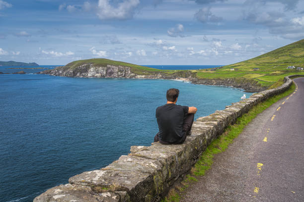 hombre de mediana edad sentado en una pared, admirando una vista increíble de slea head y la península de dingle - republic of ireland famous place dingle peninsula slea head fotografías e imágenes de stock